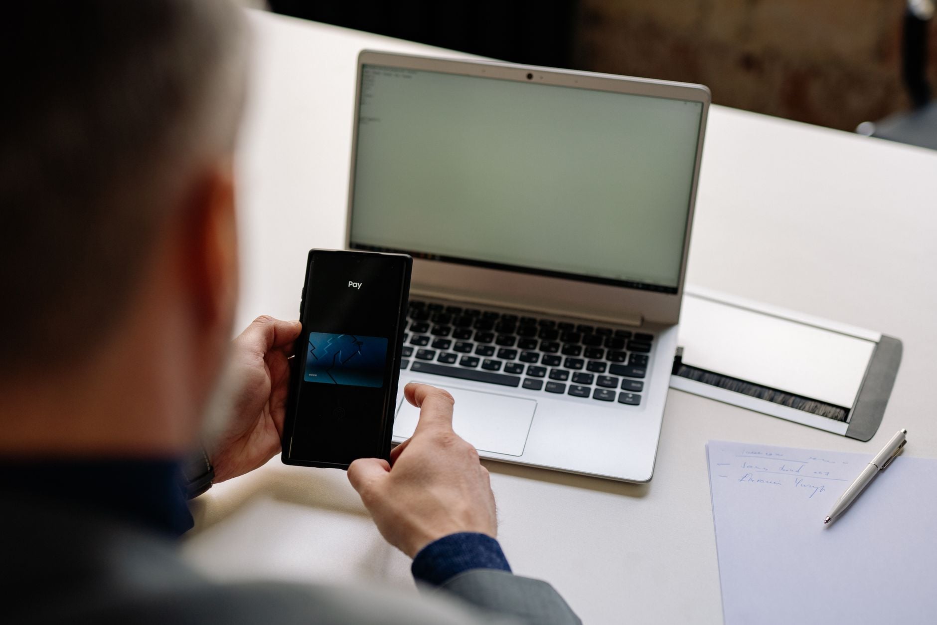 Mann hält Smartphone mit Pay-Funktion vor Laptop auf Schreibtisch