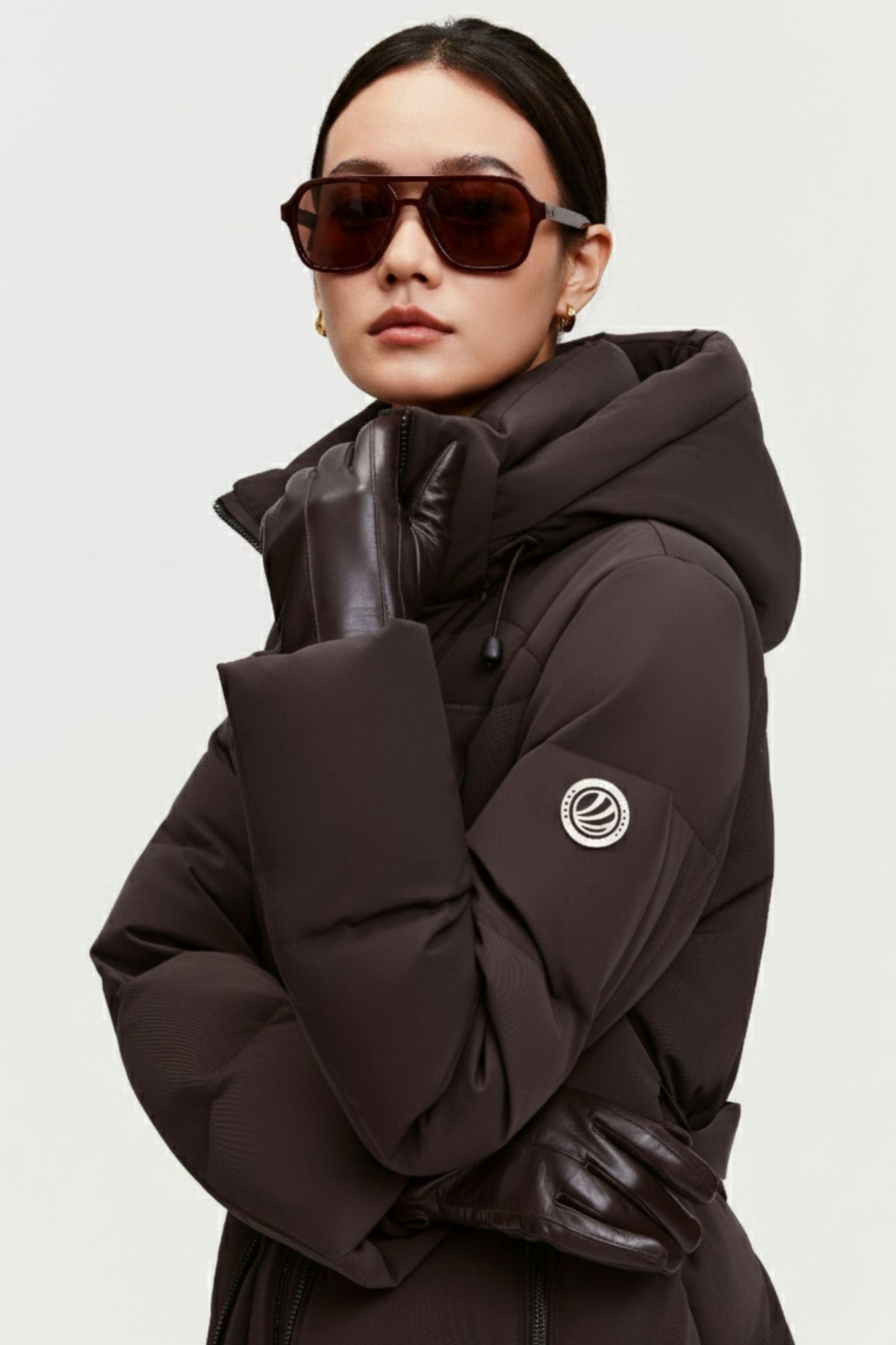 Woman wearing dark brown puffer jacket with hood and oversized fit, paired with brown aviator sunglasses