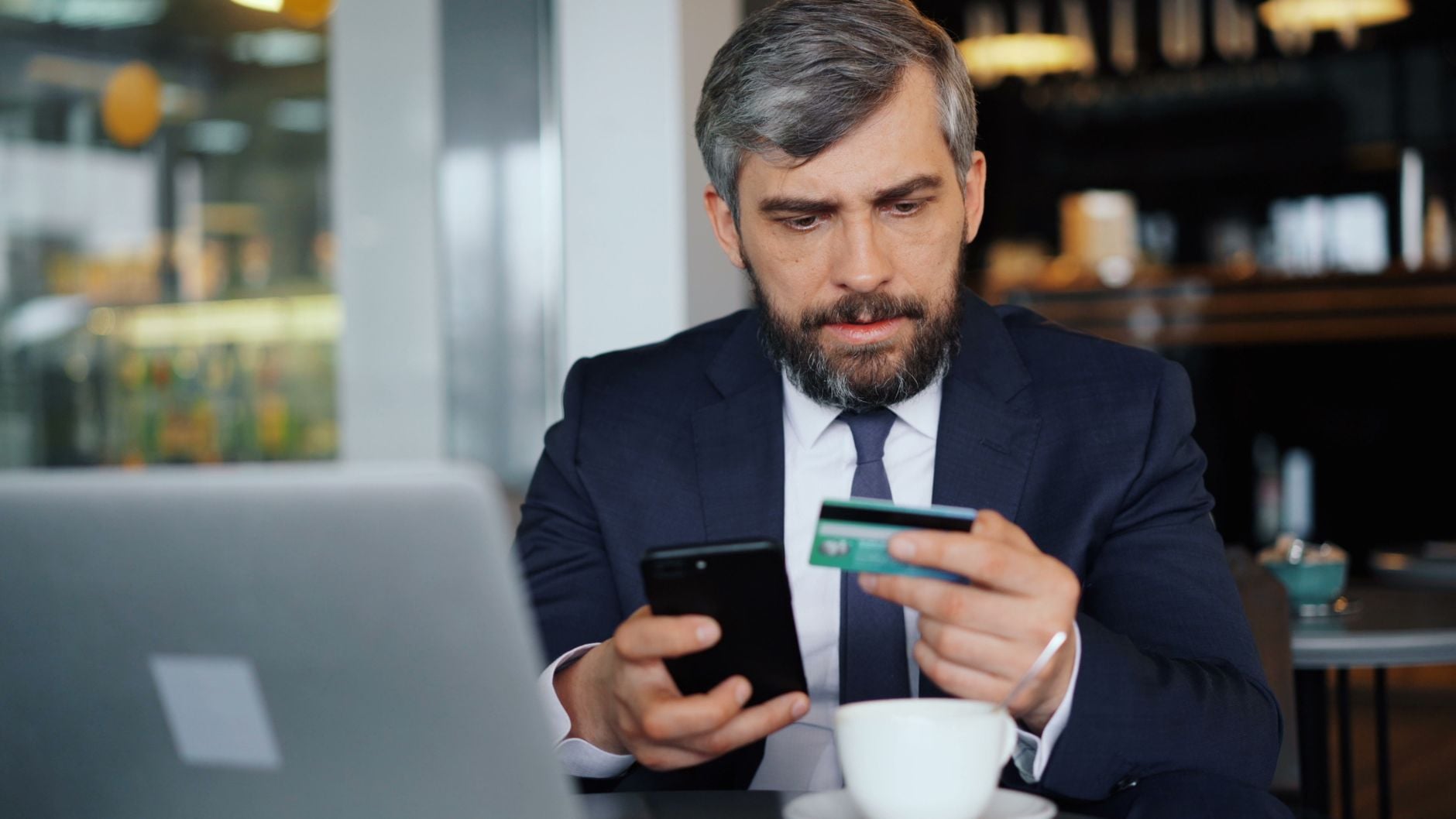 Geschäftsmann in Anzug mit Smartphone und Kreditkarte am Laptop im Büro